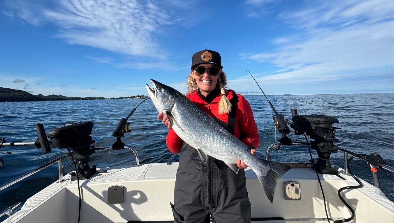 A guest in a red jacket holds a Coho salmon at the bow of an Eagle Pointe Lodge boat, blue sky and calm water behind her