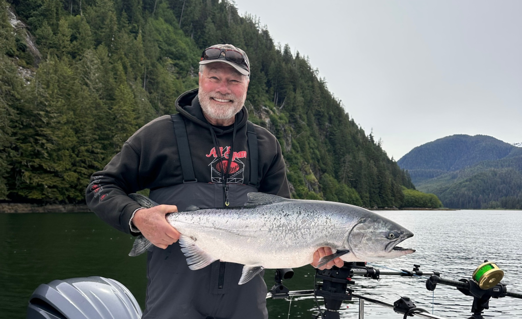 A smiling guest in a cap and fishing bibs holds a large, bright silver chinook salmon on a boat, with forested mountains and calm water behind him.
